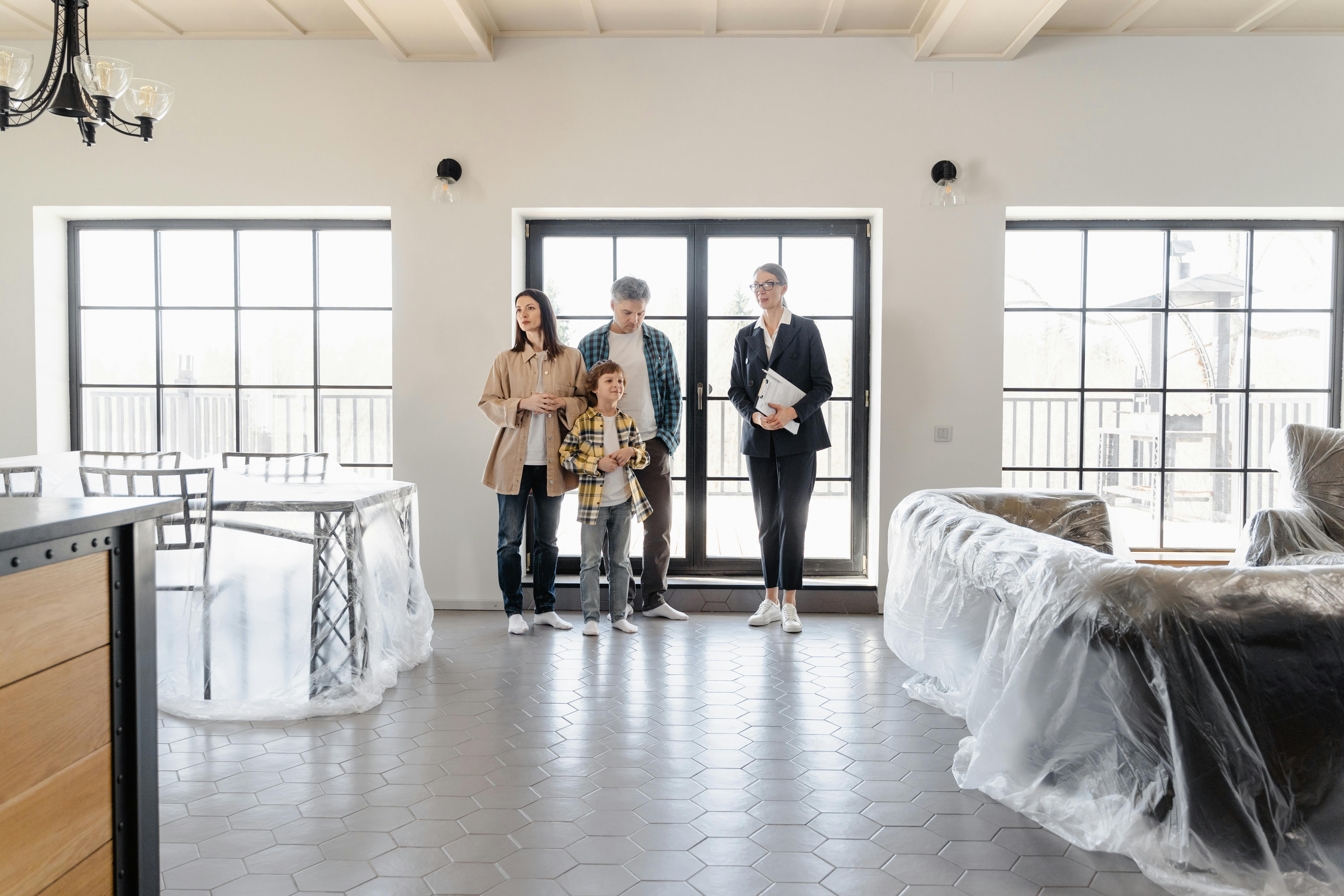 A couple and their young son touring a bright, high-windowed home with a real estate agent in a dark suit holding a clipboard, furniture around them covered in protective plastic.